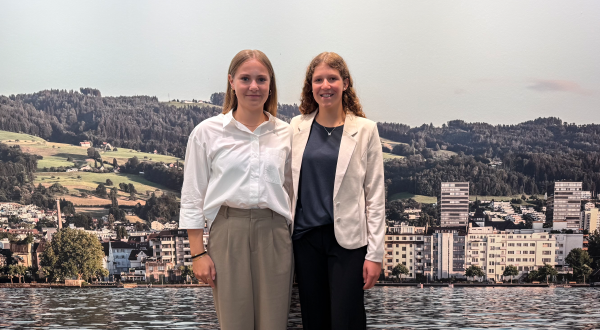 Zwei junge Frauen stehen vor einer malerischen Landschaft mit Seen und Bergen, lächelnd in formeller Kleidung.