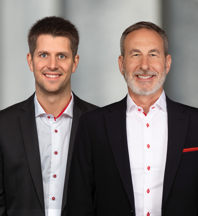 Zwei Männer in Anzügen mit Hemden mit roten Akzenten, lächelnd vor neutralem Hintergrund.