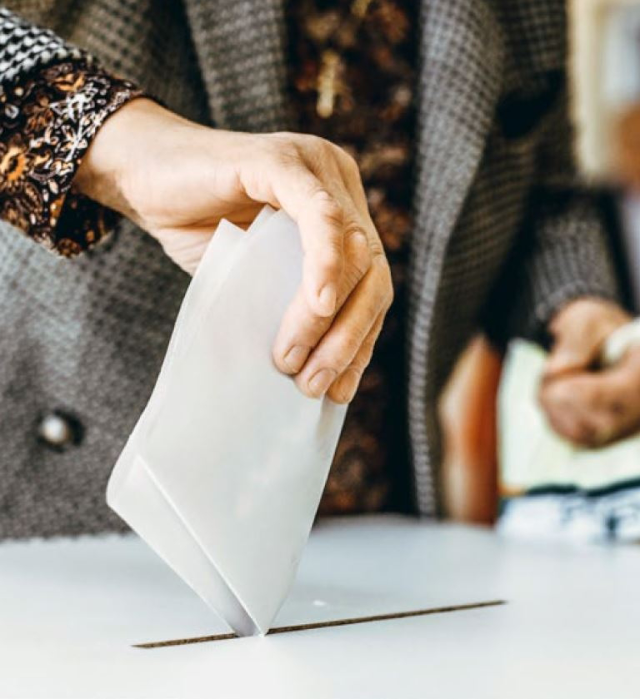 Wahlzettel wird in eine Wahlurne eingeworfen, mit Fokus auf die Hand und den Umschlag.