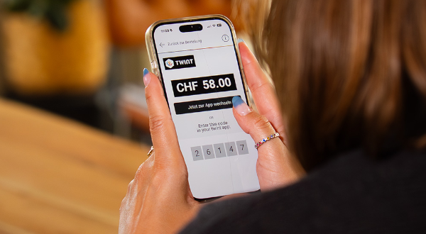 Person hält ein Smartphone und zeigt den TWINT-Bildschirm mit CHF 58.00 und Eingabefeld für den Code.