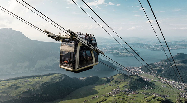 Die doppelstöckige Cabrio-Luftseilbahn schwebt mit Fahrgästen auf dem offenen Oberdeck vor einem Bergpanorama mit Blick auf den Vierwaldstättersee.