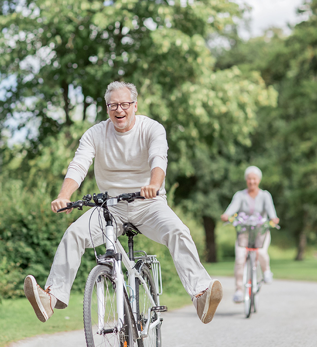 Ein älterer Mann fährt vergnügt mit ausgestreckten Beinen auf einem Fahrrad durch eine grüne Parkanlage, während ihm im Hintergrund eine Frau auf dem Velo folgt.