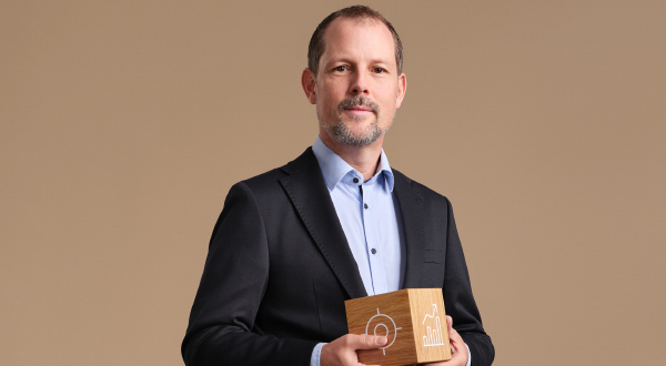 Mann in Anzug hält eine Holzfigur mit Symbolen in der Hand, vor beige Hintergrund.