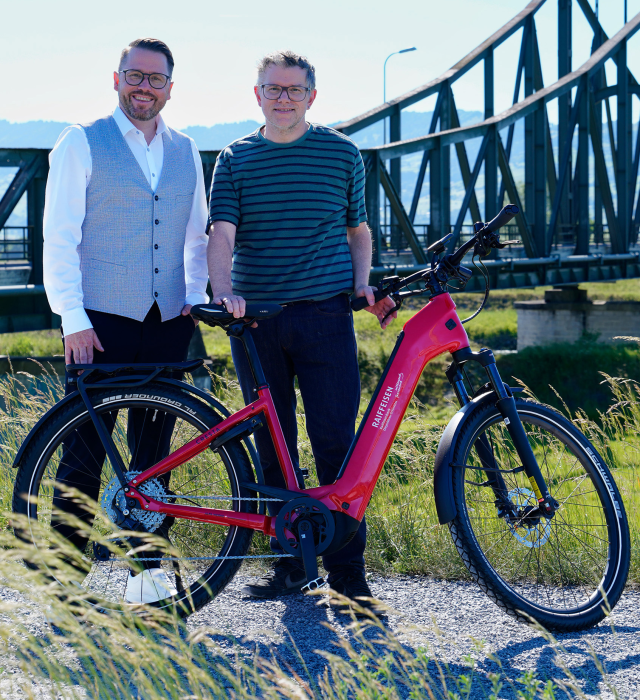 Zwei Personen posieren mit einem roten E-Bike vor einer Brücke in der Natur.