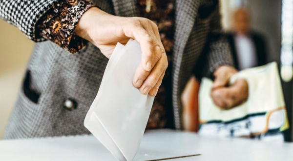 Wahlzettel wird in eine Wahlurne eingeworfen, mit Fokus auf die Hand und den Umschlag.
