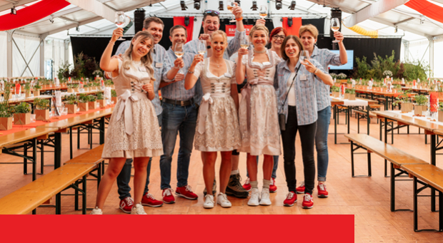 Gruppe von Personen in Trachtenkleidung steht in einem Festzelt zusammen und hebt Gläser zum Anstossen zwischen langen Holztischen.
