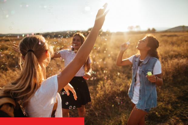 Drei junge Frauen pusten und fangen Seifenblasen auf einer Wiese im warmen Gegenlicht der Abendsonne.