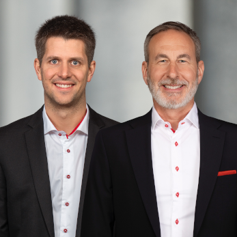 Zwei Männer in Anzügen mit Hemden mit roten Akzenten, lächelnd vor neutralem Hintergrund.