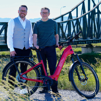 Zwei Personen posieren mit einem roten E-Bike vor einer Brücke in der Natur.