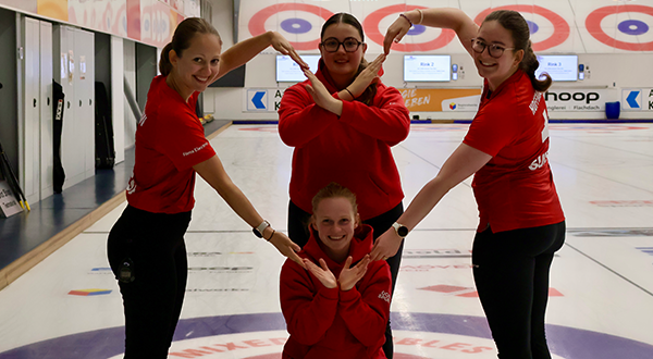 Vier lächelnde Curlerinnen in roter Teamkleidung posieren auf dem Eis einer Curlinghalle und formen mit ihren Armen gemeinsam ein grosses Herz.