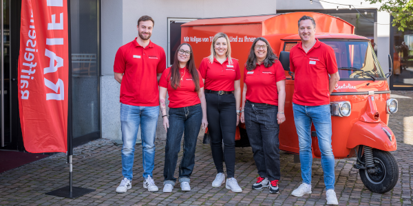 Teamfoto mit fünf Personen in roten Shirts vor einem orangenen Auto und einer Raiffeisen Werbefahne.