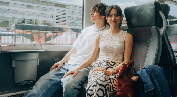 Junges Paar im Zug, entspannt sitzend mit sportlicher Kleidung, einer mit einer roten Tasche.