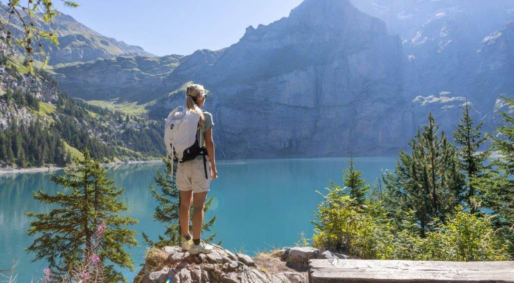 Person mit Rucksack steht auf einem Felsen und blickt über einen türkisfarbenen Bergsee in alpiner Landschaft.