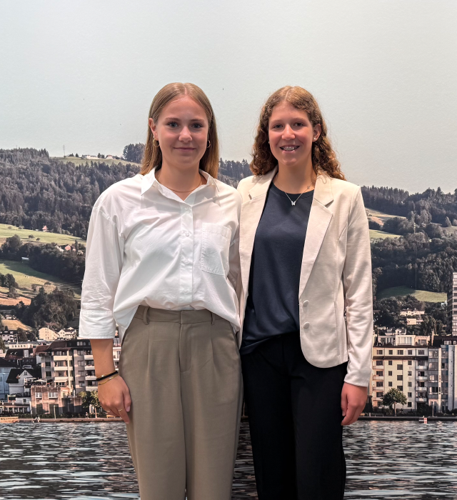 Zwei junge Frauen stehen vor einer malerischen Landschaft mit Seen und Bergen, lächelnd in formeller Kleidung.
