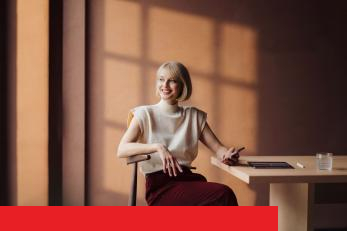 Eine stilvoll gekleidete Frau mit blondem Bob sitzt lächelnd an einem modernen Besprechungstisch, hält ihr Smartphone und blickt zur Seite ins einfallende Sonnenlicht.
